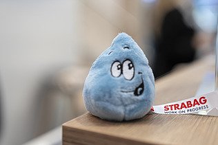 Close-up of a friendly, blue water droplet plush toy with a STRABAG ribbon, serving as a charming giveaway.