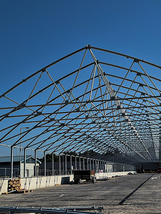 Construction phase of a solar sludge drying facility showing a large open concrete base with a partially assembled steel frame structure under a clear blue sky.